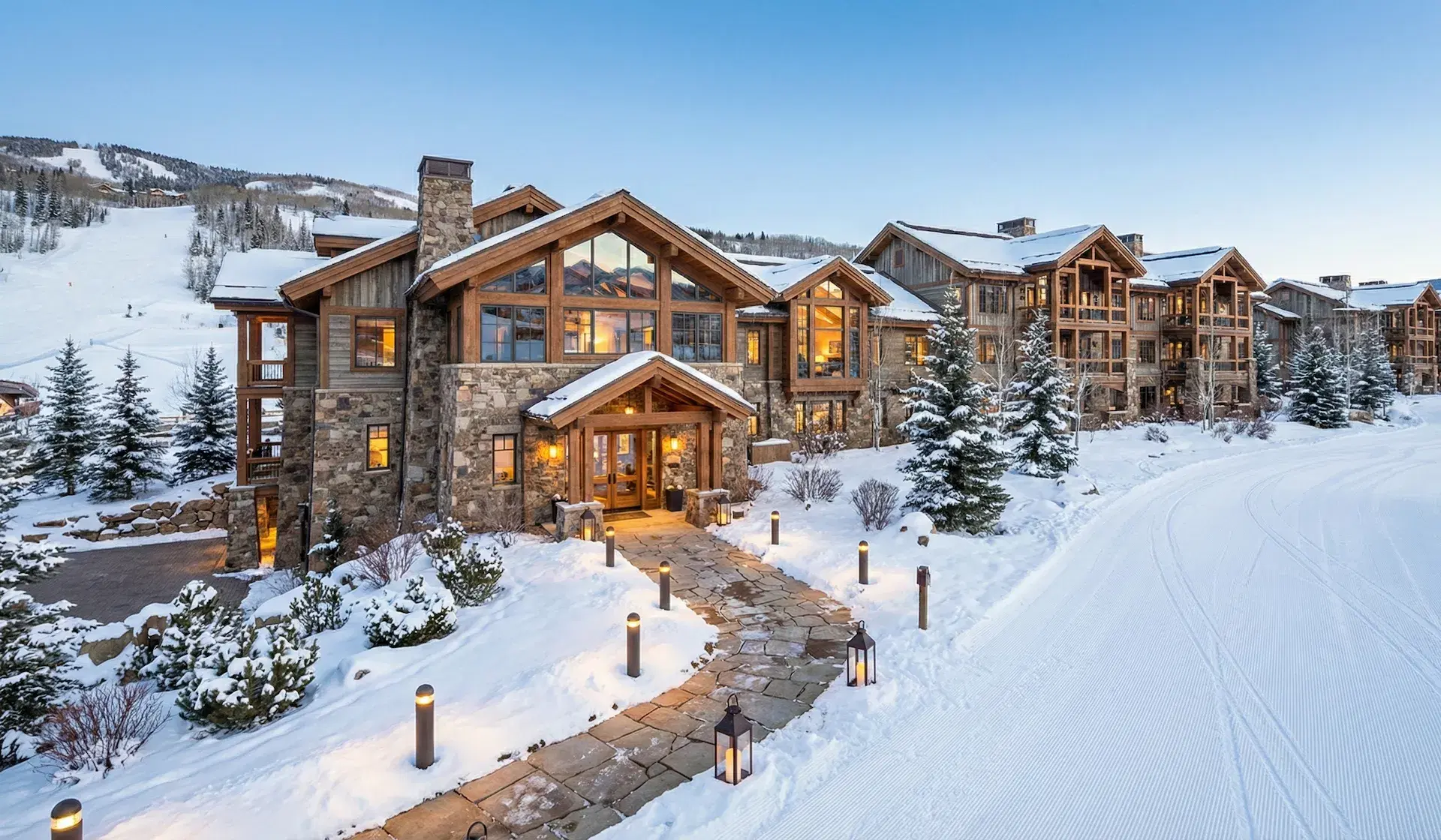 Mountain lodge at dusk with snow-covered roofs, glowing windows, and a lantern-lined path beside a groomed ski run